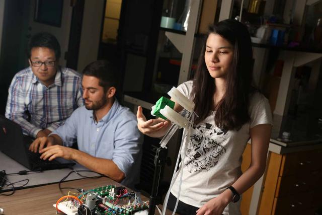 Ph.D. students Benjamin Shih and Dylan Drotman monitor a computer while Ph.D. student Adriane Minori gets ready to remove a 3D-printed object from the soft robotic gripper.
